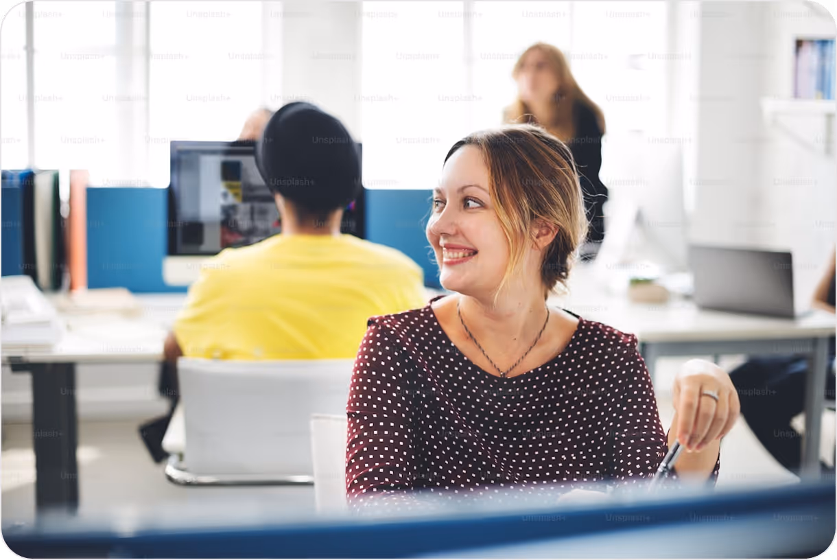 Smiling woman in a polka dot top looking sideways in a bright office with coworkers working at desks in the background.