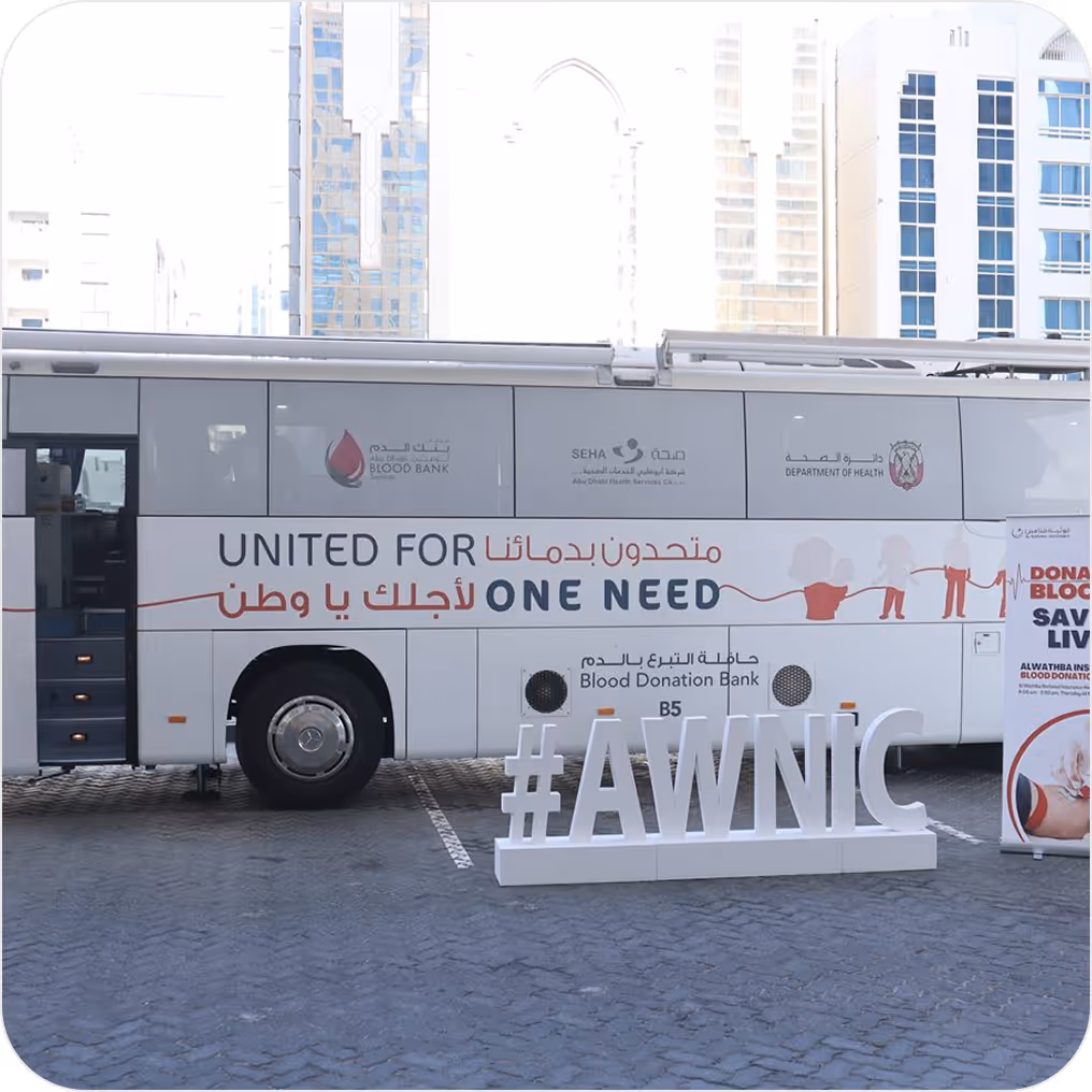 Side view of a blood donation bus with the text 'UNITED FOR ONE NEED' in English and Arabic, and a hashtag sign #AWNIC in front of it.