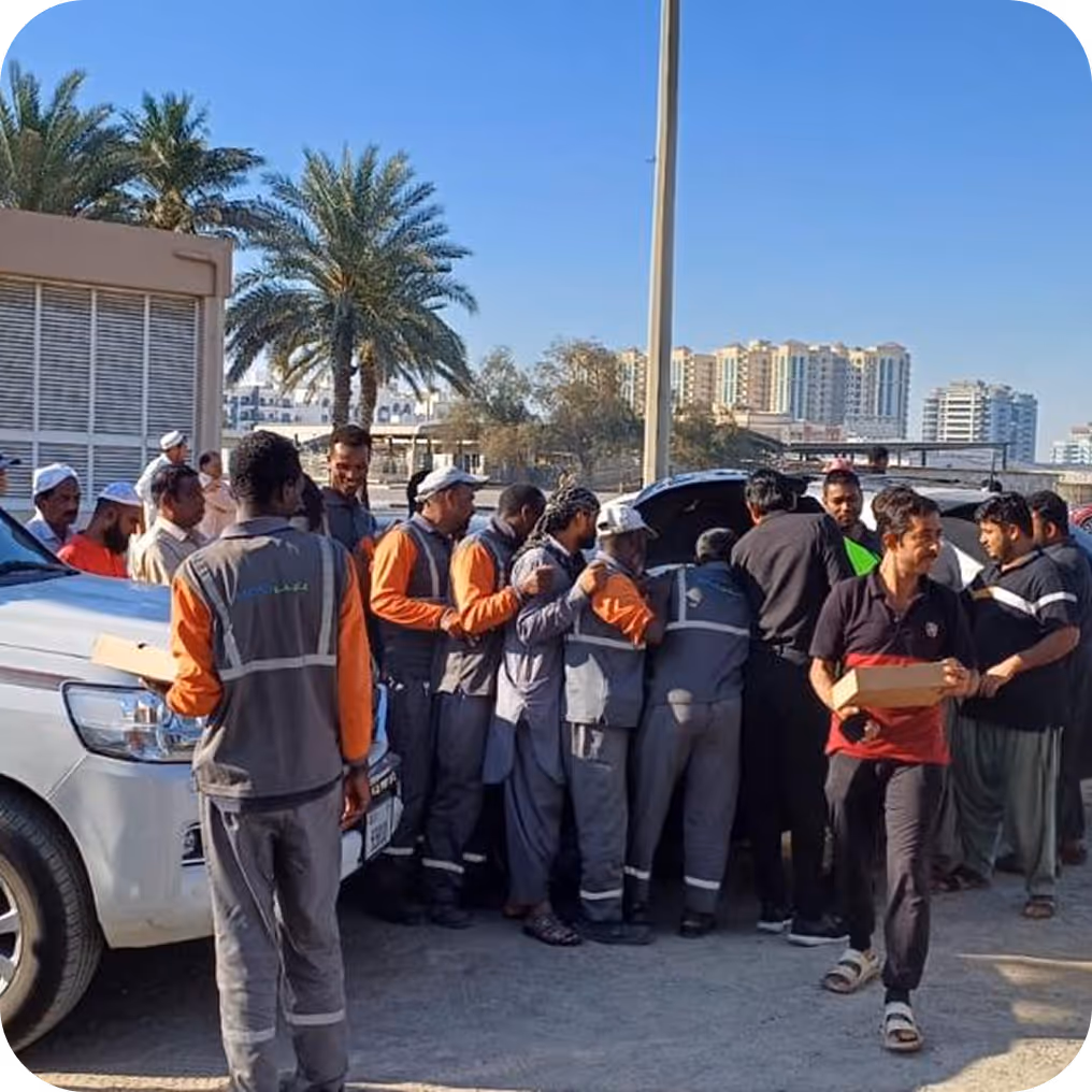Group of men, many in work uniforms, gathering around vehicles outdoors under clear sky, some holding boxes.