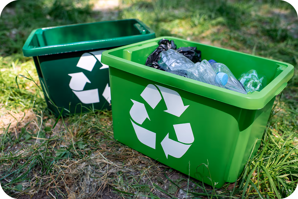 Two green recycling bins on grass, one filled with plastic bottles and bags, both displaying white recycling symbols.