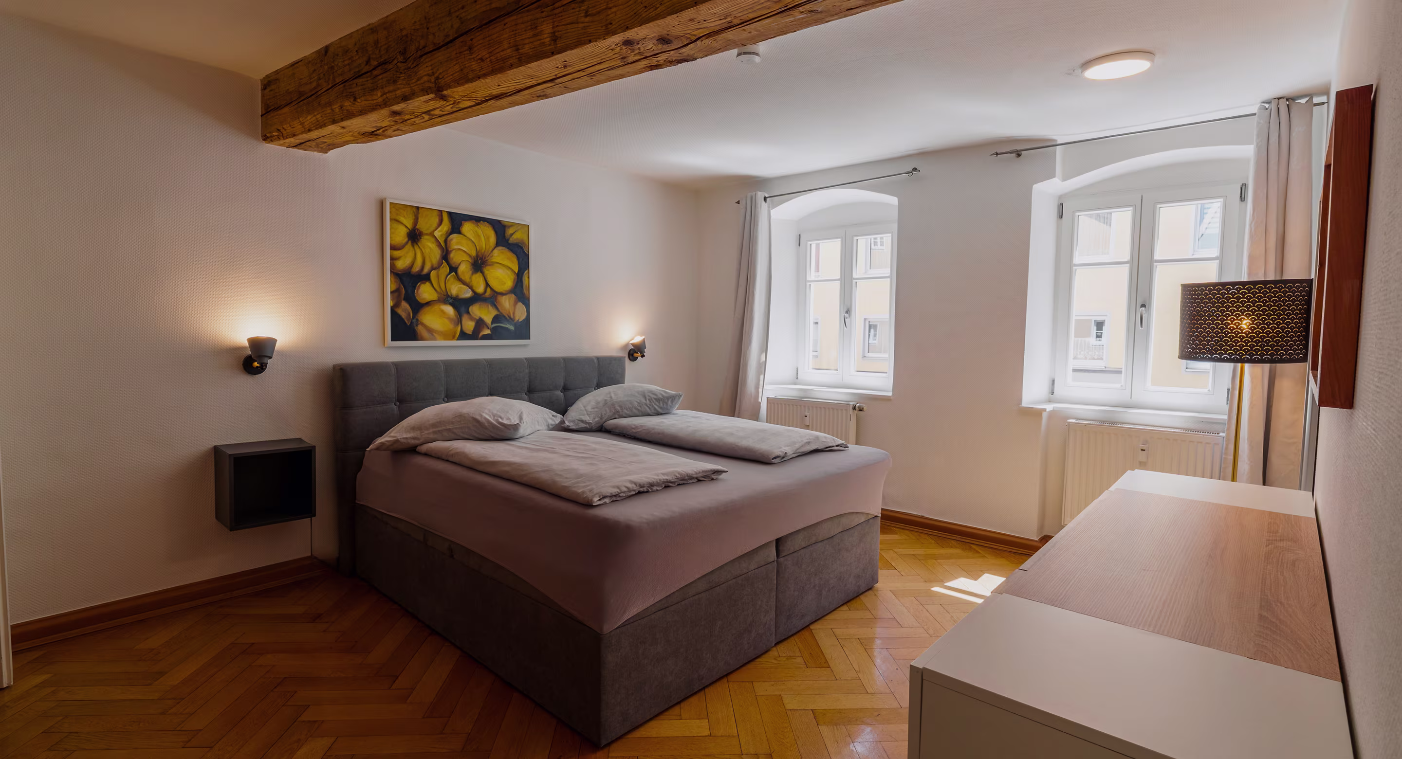 Cozy bedroom with double bed, wooden floor, yellow flower painting, and two windows with curtains.