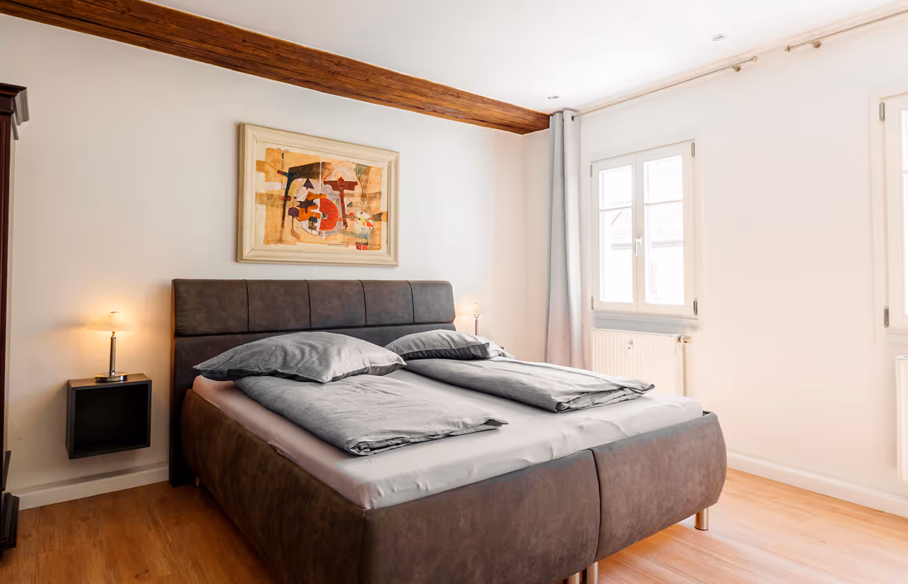 Modern bedroom with a brown upholstered bed, gray bedding, wooden floor, and abstract painting on white wall.