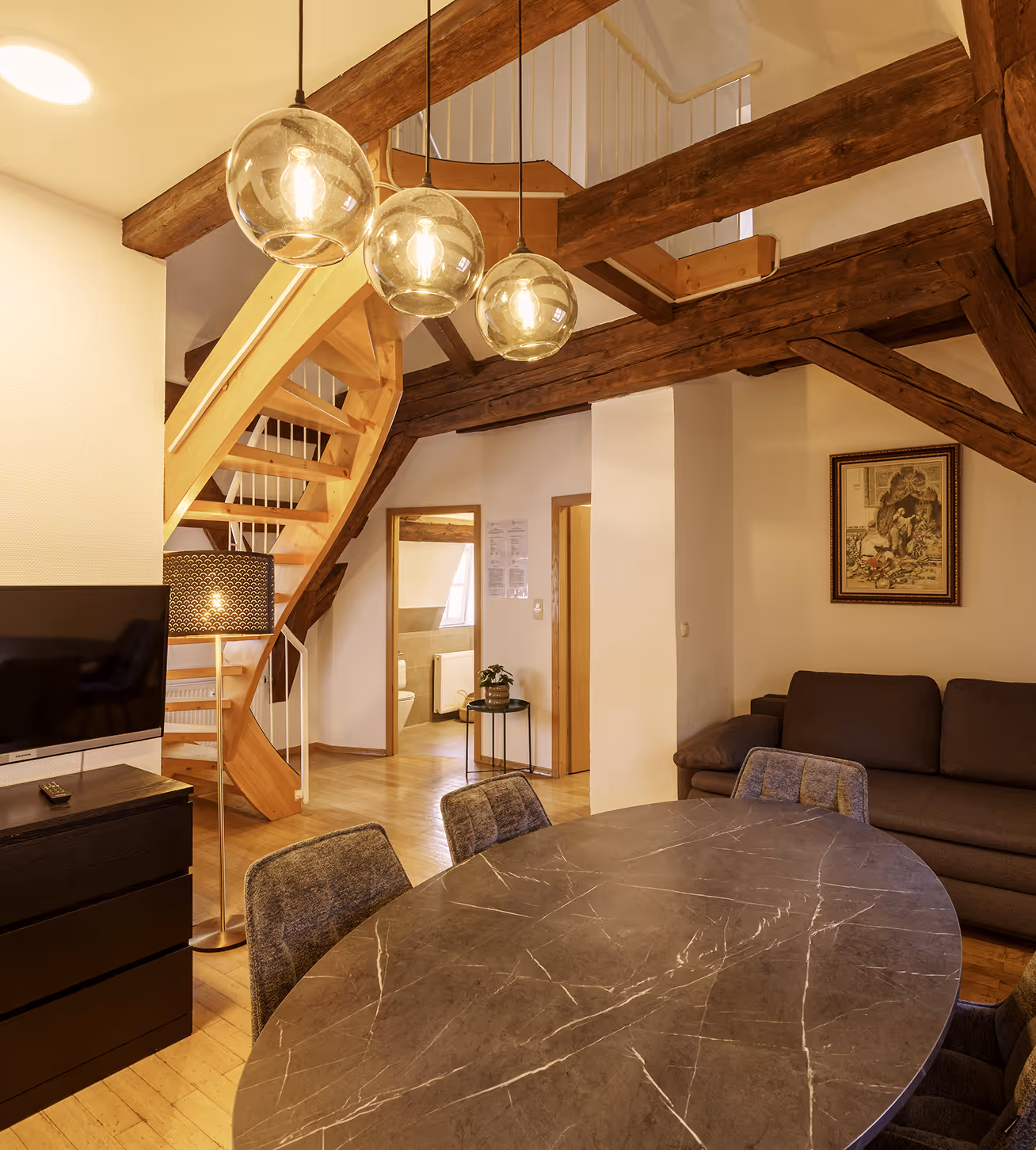 Cozy living space with wooden beams, round marble table, gray chairs, sofa, and a staircase with hanging lights.