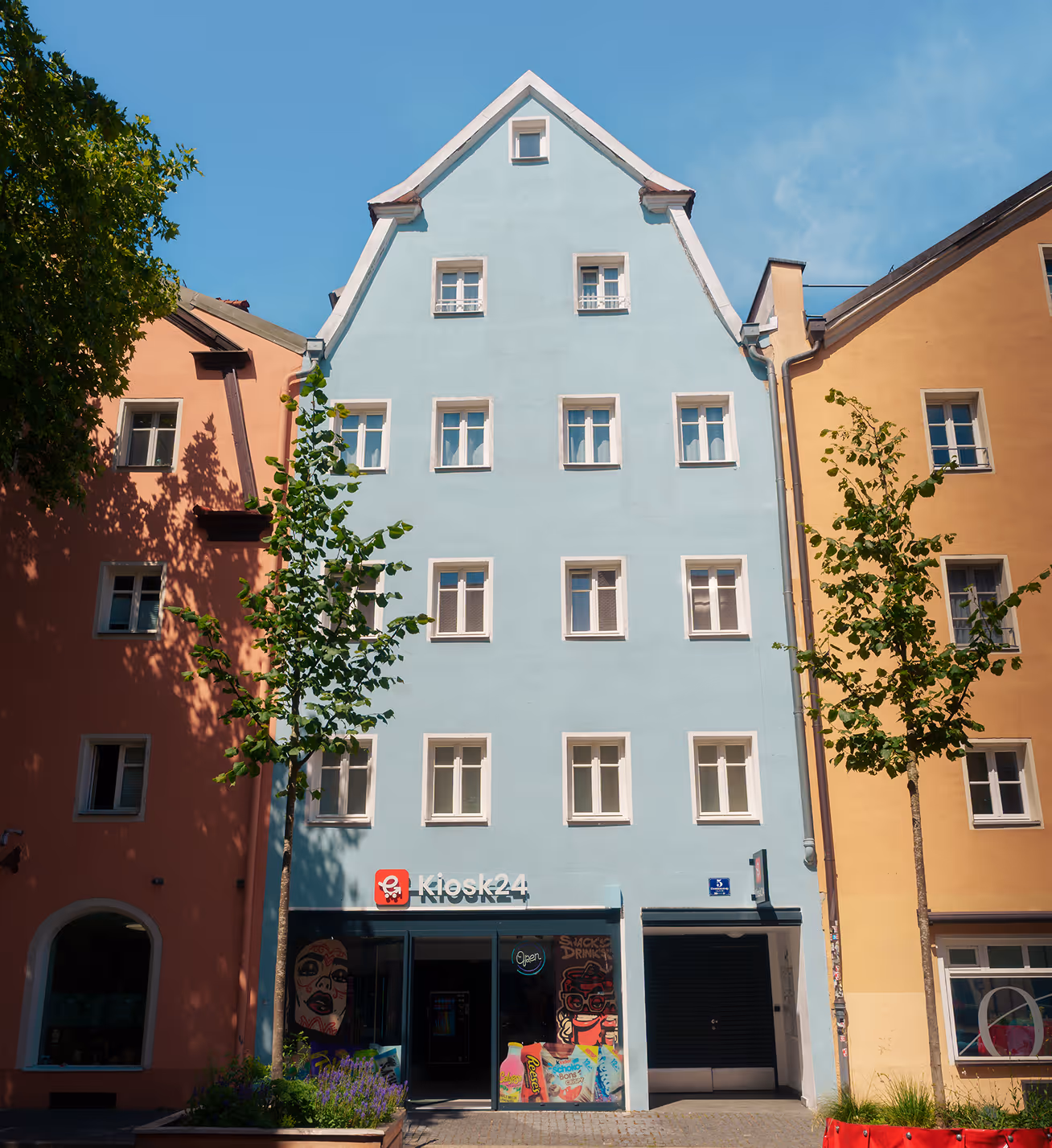 Blue building with multiple windows and kiosk24 store on ground floor under a clear sky.
