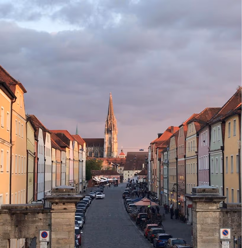 Cobblestone street lined with colorful buildings leading to a Gothic cathedral under a cloudy sky.