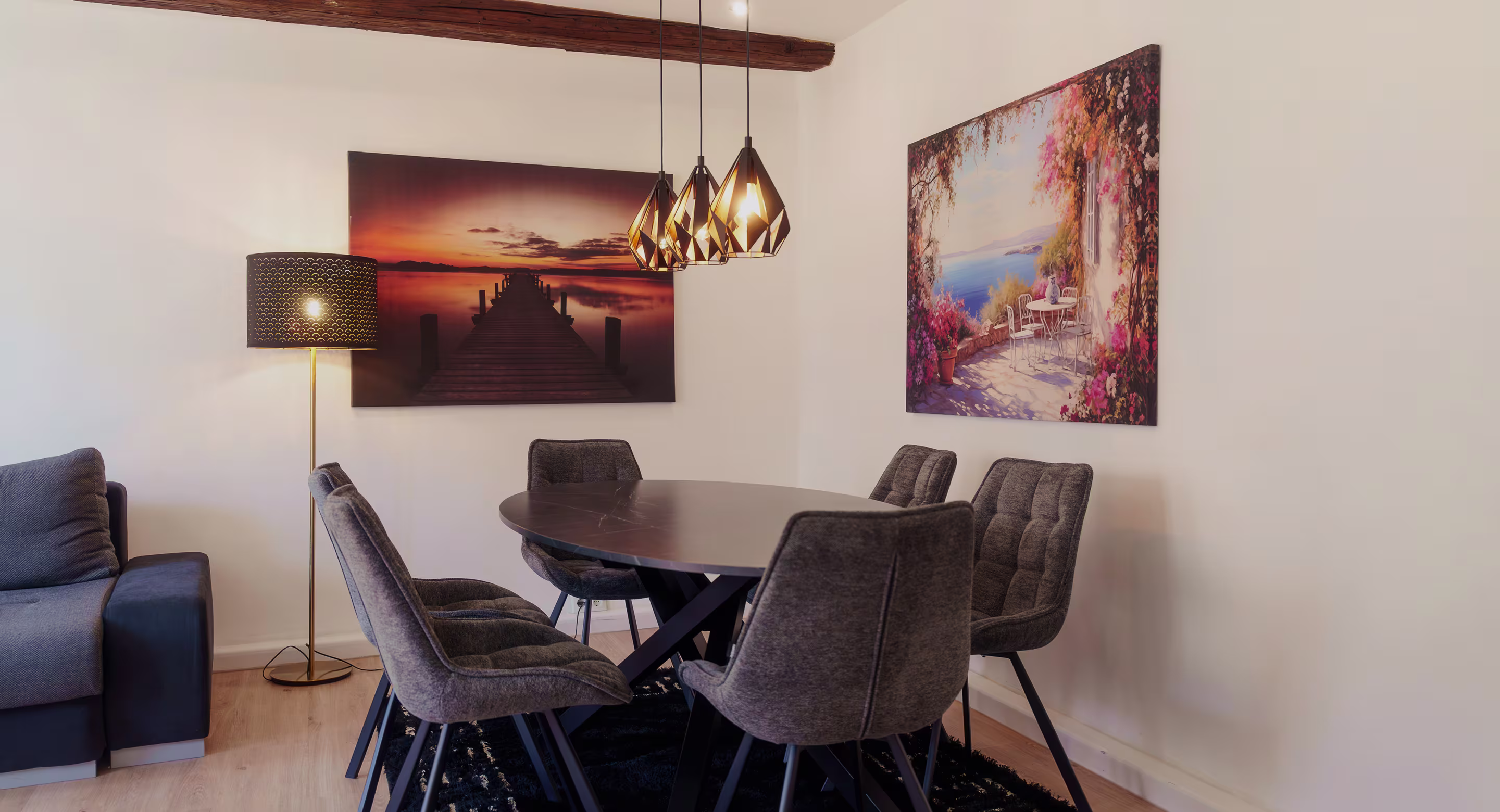 Dining area with a round table, six cushioned chairs, a floor lamp, and two wall paintings.