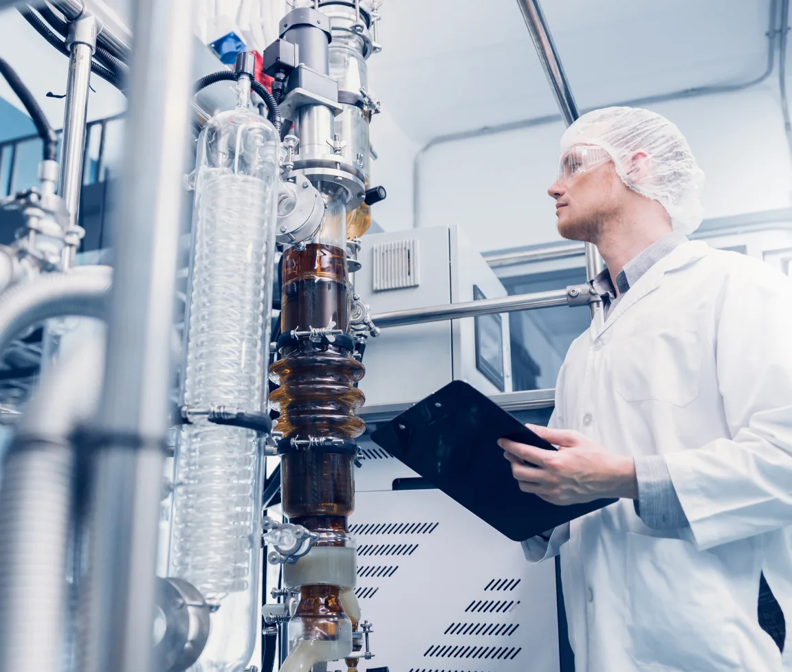 Laborant mit Schutzbrille und Kittel überprüft Anlagen in einem Labor mit transparenten Rohren.