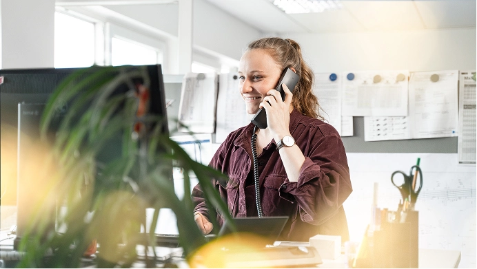 Frau spricht lächelnd am Telefon und arbeitet an einem Computerschreibtisch im Büro.