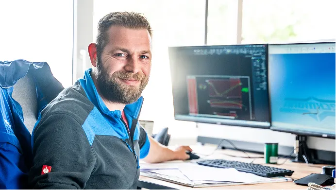 Ein lächelnder Mann sitzt vor zwei Monitoren mit technischen Zeichnungen am Schreibtisch.