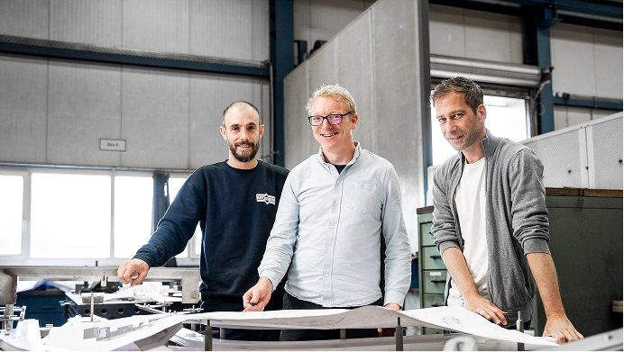 Drei Männer in einer Werkstatt, die zusammen an technischen Zeichnungen arbeiten.