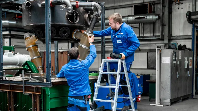 Zwei Männer in blauen Overalls warten an einem Industriegerät, einer steht auf einer Leiter.