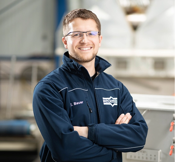 Lächelnder Mann mit Brille in blauer Arbeitsjacke steht mit verschränkten Armen in einer Fabrikhalle.