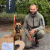 Man squatting on a path outdoors with two German Shepherd dogs sitting on either side.