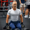 Smiling man in white shirt lifting dumbbells while sitting in a gym with other people in background.