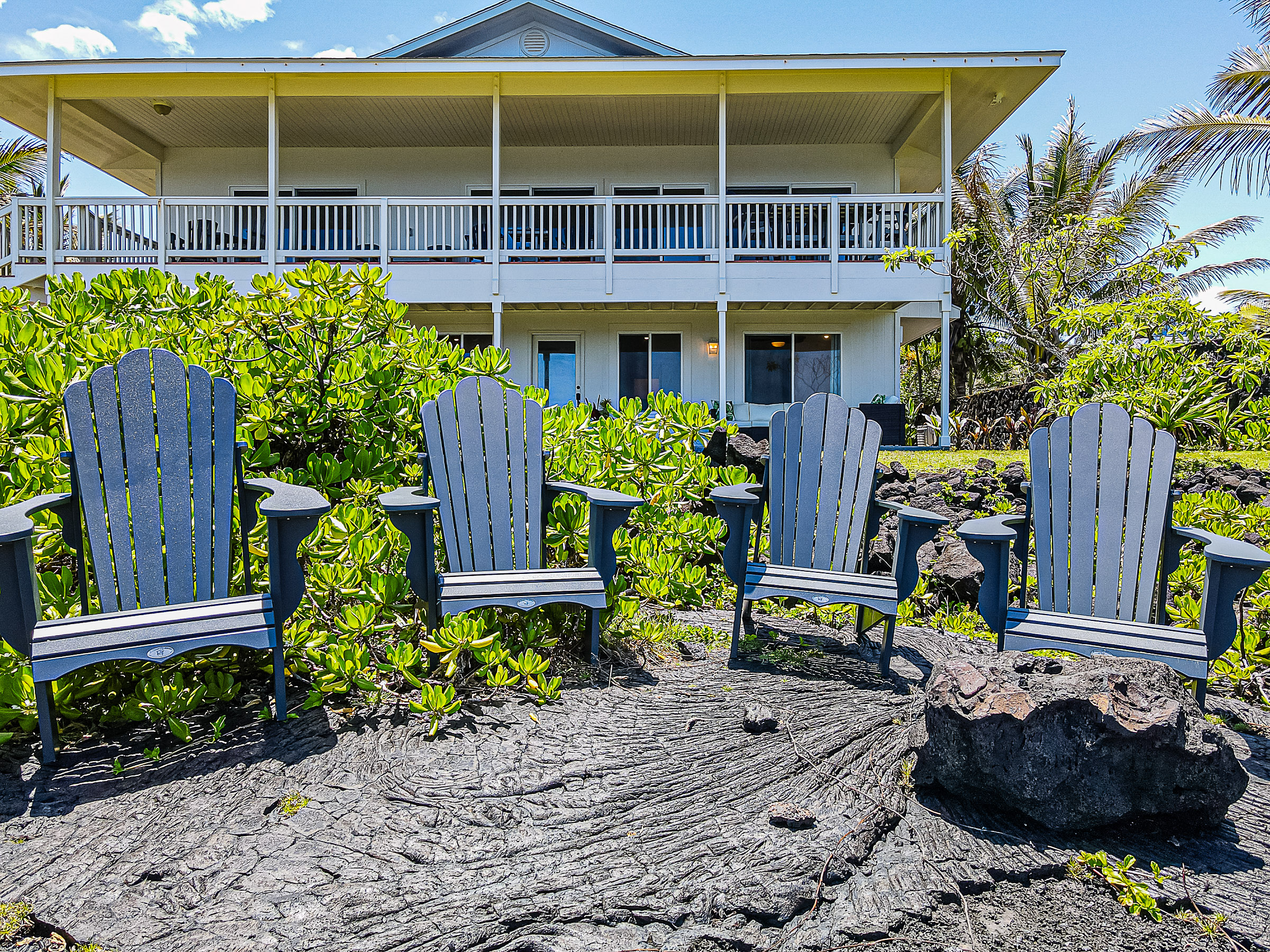 Four empty blue Adirondack chairs arranged on volcanic rock with a two-story white house and lush greenery in the background.