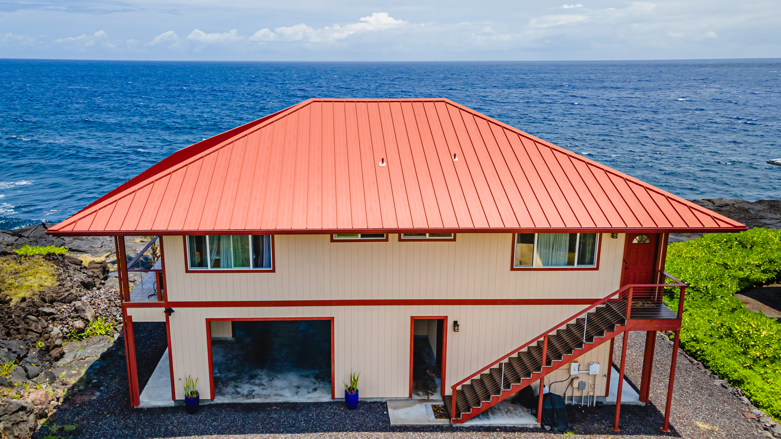 Two-story beige house with red trim and roof overlooking the ocean, surrounded by rocky terrain and greenery.