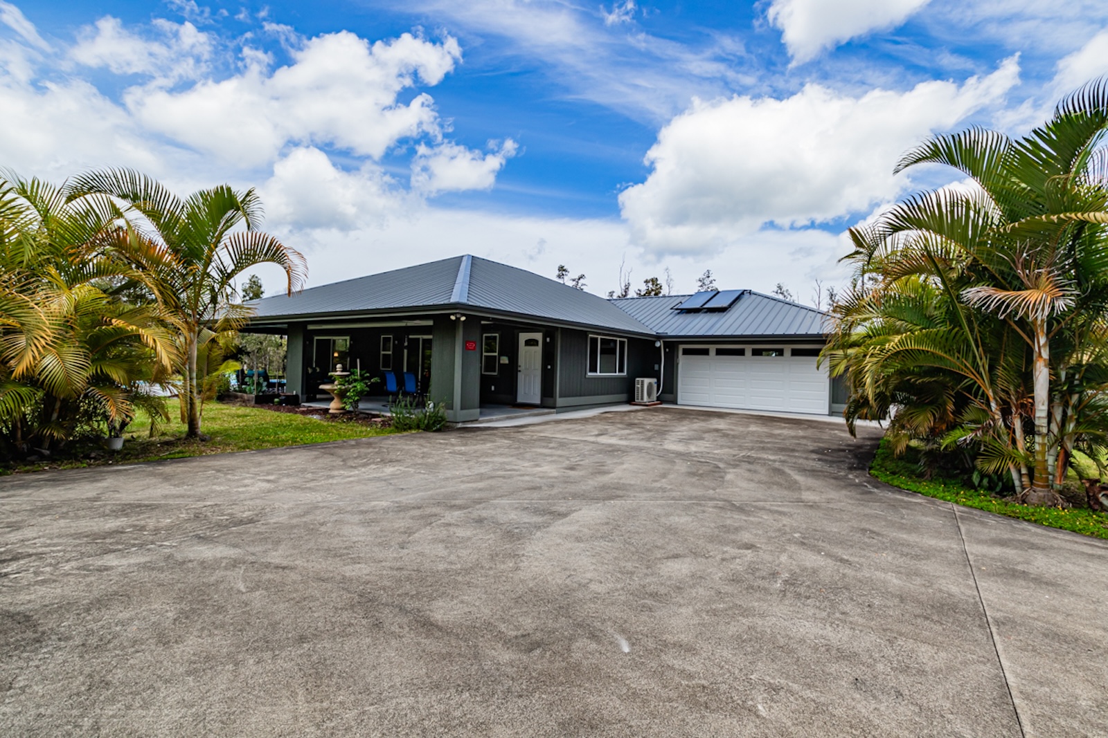 Single-story house with grey metal roof, white garage door, and large driveway bordered by palm trees under a blue partly cloudy sky.