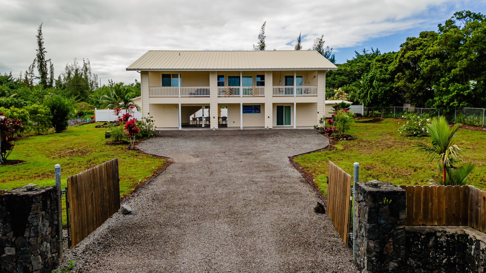 Two-story beige house with a metal roof, open front yard with gravel driveway, green lawn, and tropical plants on the sides.
