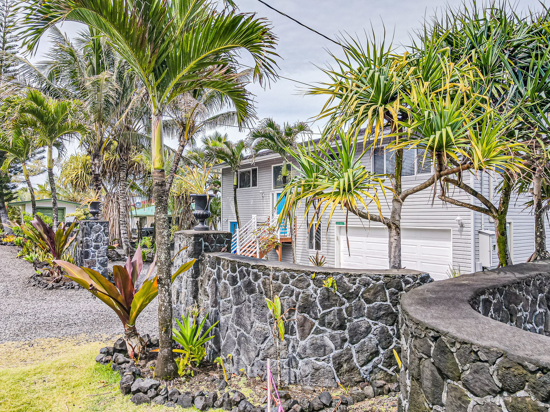 Modern gray house behind a decorative stone wall with tropical palm trees and plants in the yard.