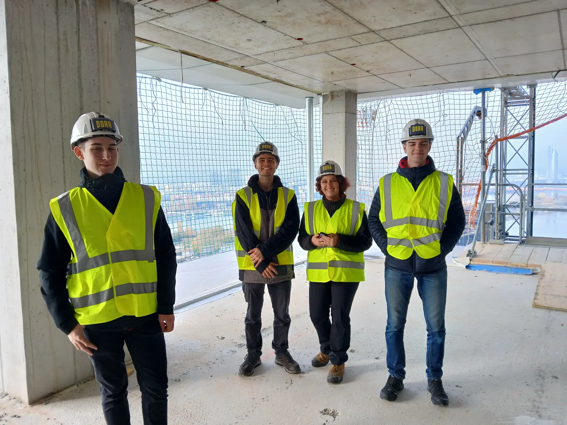Čtyři lidé v reflexních vestách a přilbách stojí na staveništi s panoramatem města za bezpečnostní sítí.
