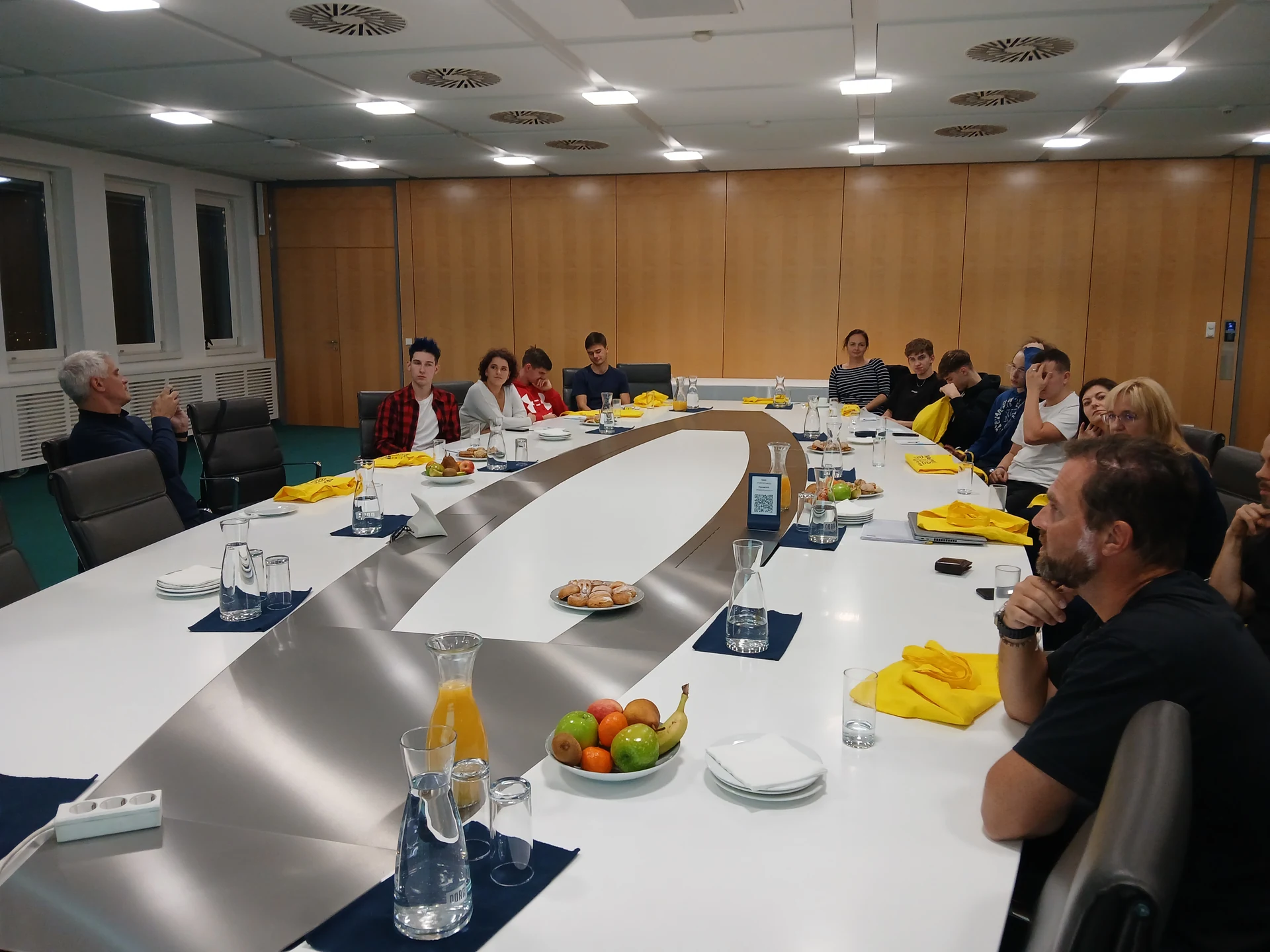 Skupina lidí sedící kolem oválného konferenčního stolu s džbány vody a ovocem.