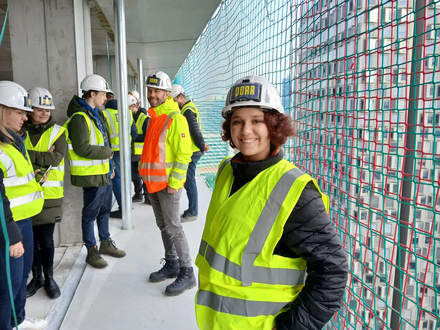 Skupina lidí s bezpečnostními helmami a reflexními vestami na staveništi budovy.