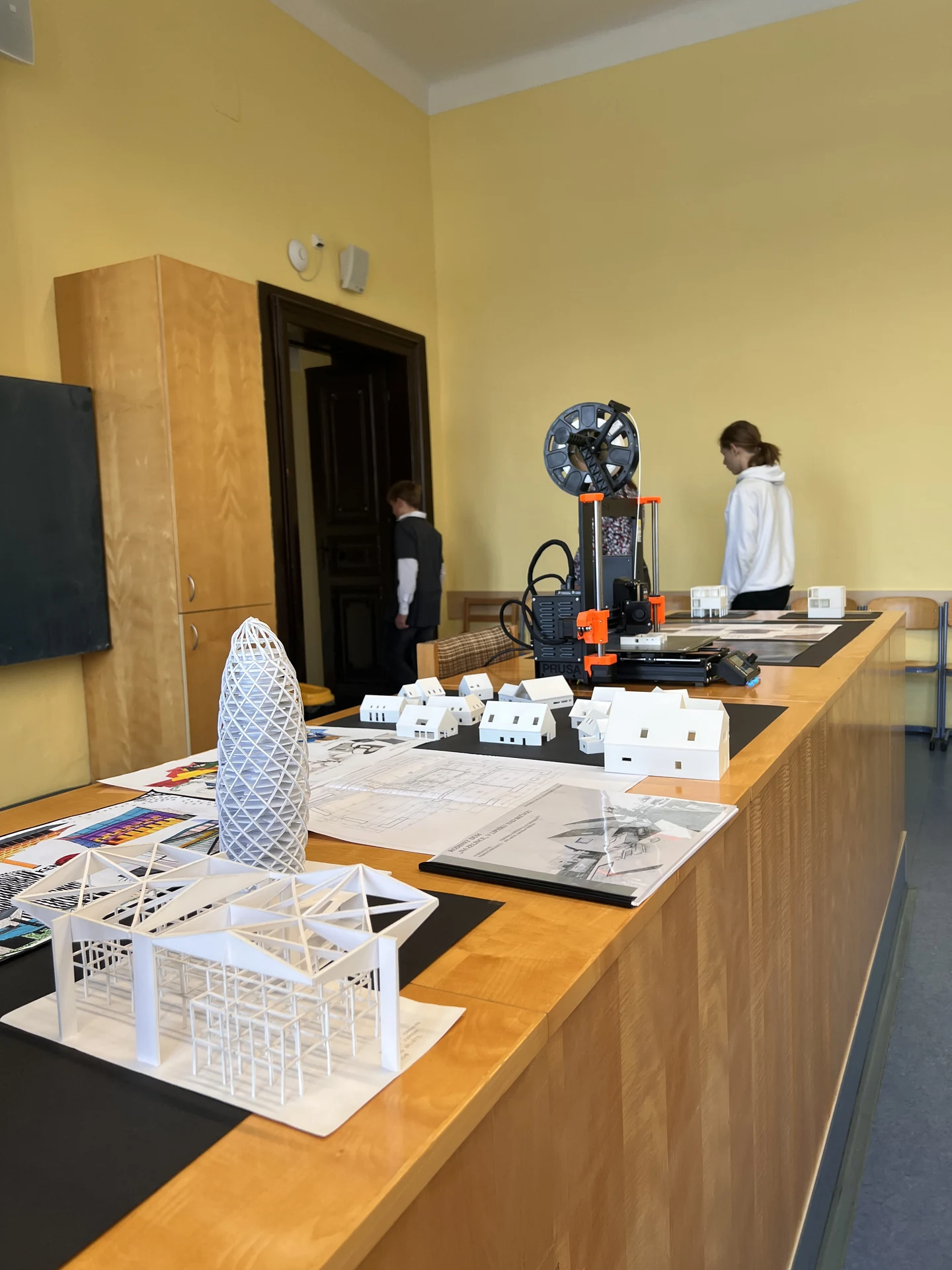 Modely bílých architektonických staveb a 3D tiskárna na dřevěném stole v učebně.