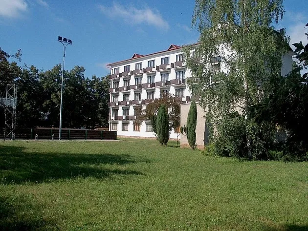 Bílá budova s mnoha okny za zeleným travnatým prostorem a stromy na modré obloze.