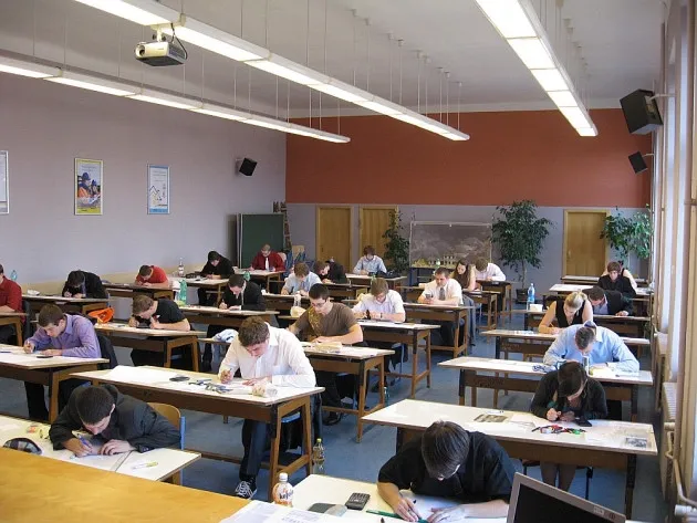 Studenti píší test v posluchárně se stoly a projektorem na stropě.