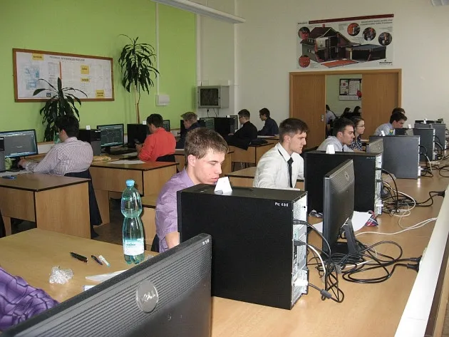 Skupina mužů pracujících u stolů s počítači v učebně s jasnými stěnami a rostlinami.