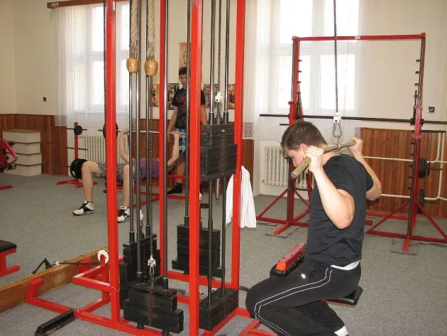 Muž cvičí s činkou na posilovací lavici, v pozadí další lidé tráví cvičení ve fitness centru.