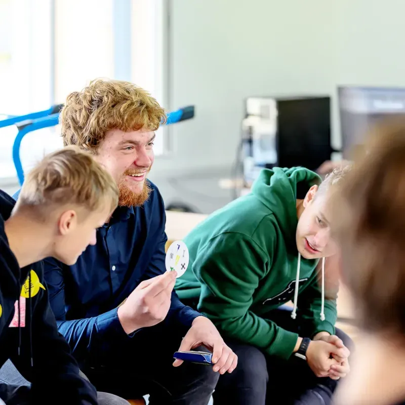 Skupina studentů sedí ve třídě a diskutuje, jeden drží kruhovou kartičku s obrázky.