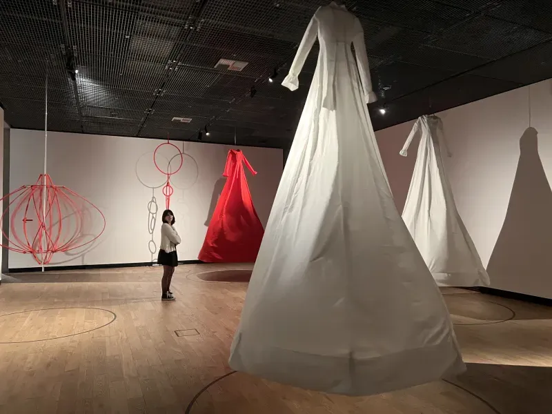Expozice textilních instalací: visící bílé a červené oděvy a kruhy ve výstavní síni s dřevěnou podlahou.