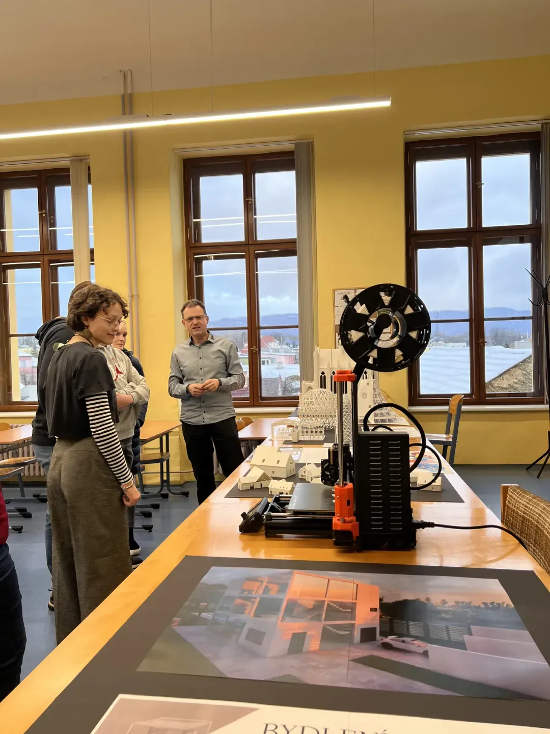 Lidé v ateliéru sledují stolní 3D tiskárnu a modely architektury na stole.