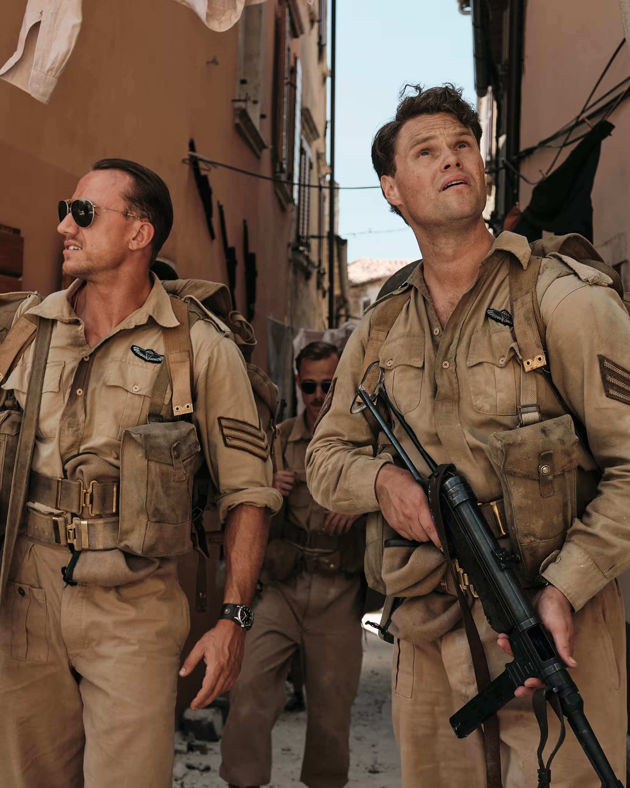 Three soldiers in tan uniforms walking through a narrow alley, one holding a rifle and looking up.