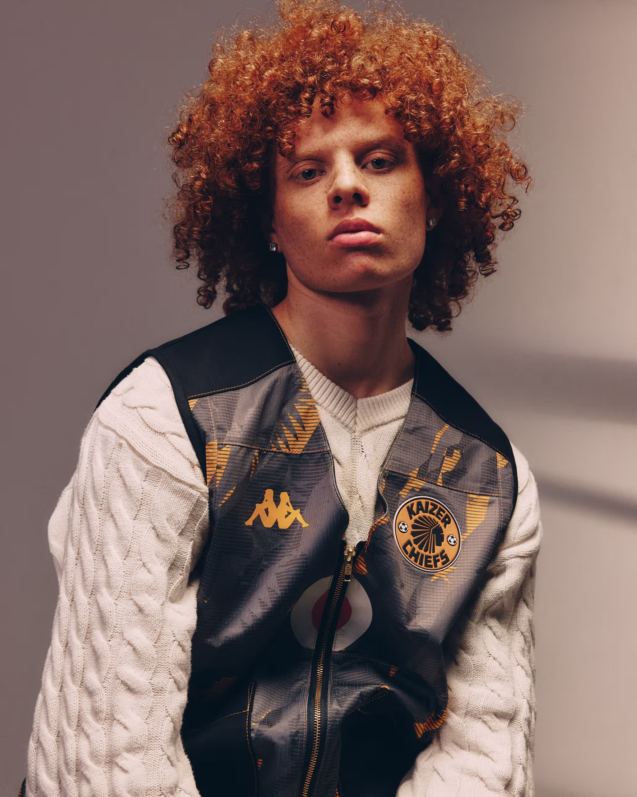 Young person with curly red hair wearing a Kaizer Chiefs jacket over a white cable knit sweater.