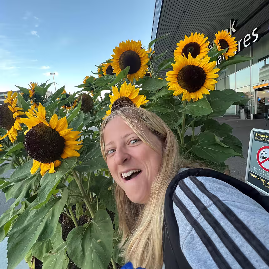 Woman with long blonde hair smiling near bright sunflowers outside a modern building.