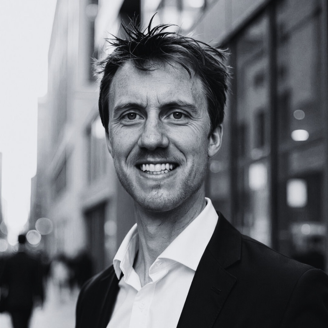 Smiling man in a suit and white shirt standing on a city street with blurred buildings behind him.