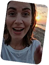Close-up of a woman with wet hair smiling and speaking, with a sunset over a rocky shore in the background.
