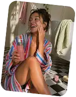 Smiling woman in a striped bathrobe sitting on a bathroom floor holding a pink smoothie cup with a straw.