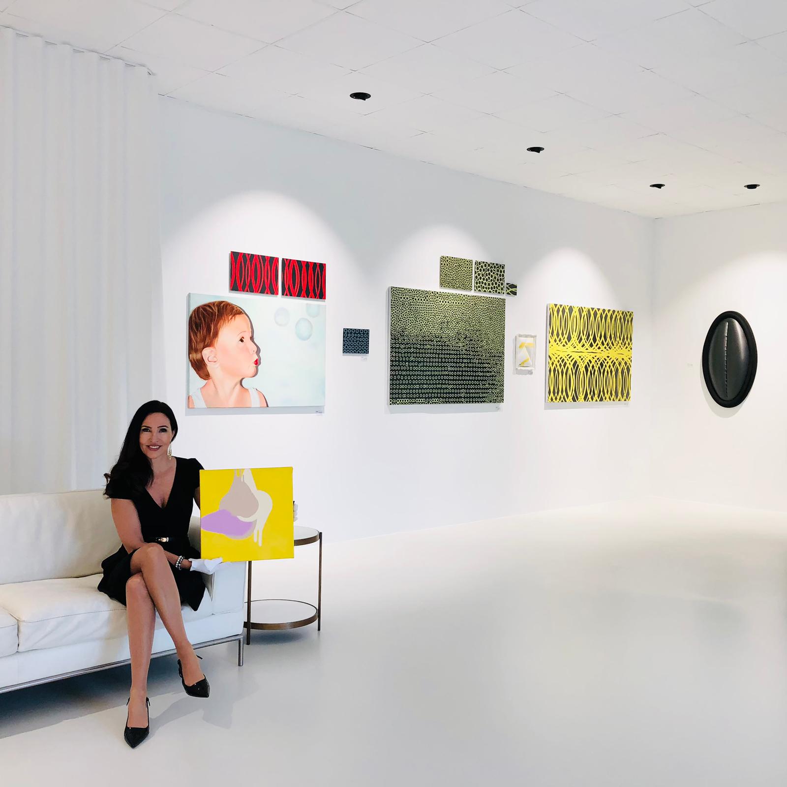 Frau in schwarzem Kleid sitzt auf weißem Sofa und hält gelbes abstraktes Gemälde in einer Galerie.