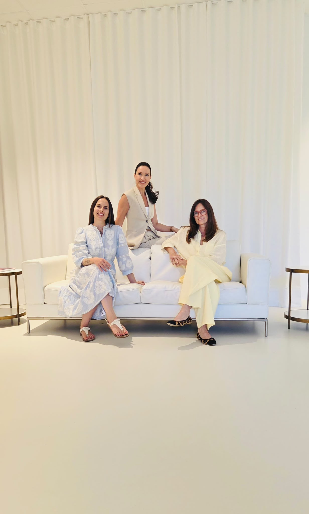 Drei Frauen, eine in blauem Kleid links, eine mittig im beigen Anzug, eine rechts in gelbem Outfit auf weißem Sofa.