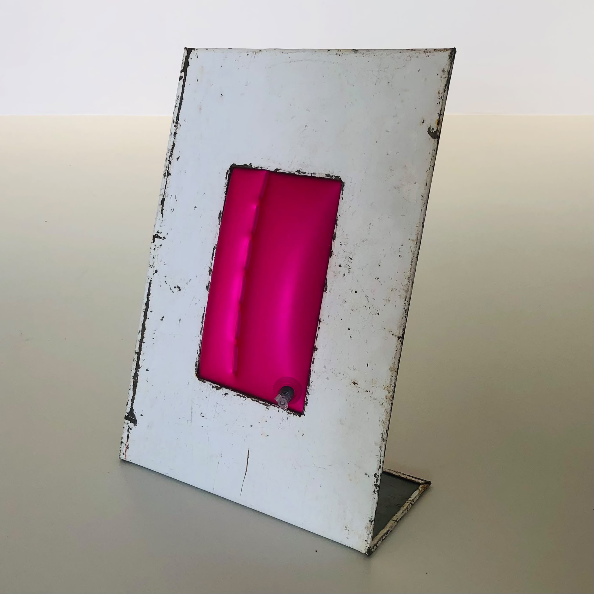 Weißes, schräg stehendes Metallblech mit einem rechteckigen Ausschnitt, darin ein pinkes aufgeblasenes Objekt.