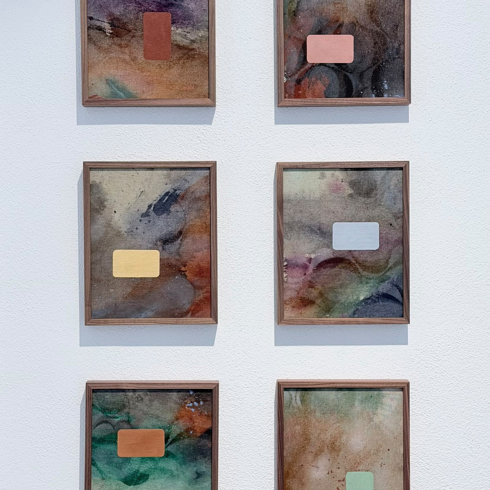 Sechs gerahmte abstrakte Gemälde auf einer weißen Wand, jedes mit einem kleinen farbigen rechteckigen Element in der Bildmitte und verschiedenen Farbmischungen im Hintergrund in der PARK GALERIE Buchkirchen bei Wels von Petuscha (Peti) Schopper.