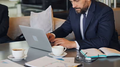 BBC - Homme en costume travaillant sur un ordinateur portable, café et documents sur la table.