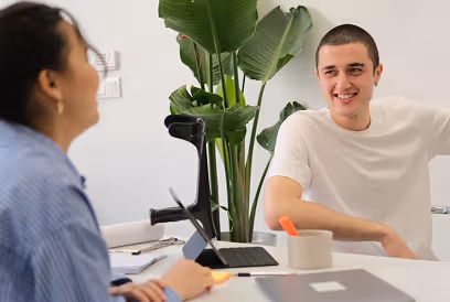 BBC - Deux personnes souriantes discutent à une table avec ordinateur et plant vert en arrière-plan.