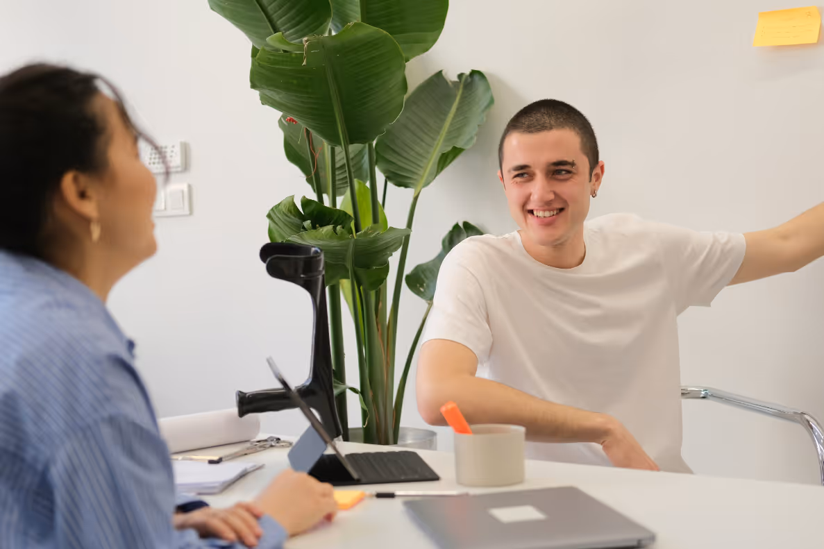 Un homme souriant en t-shirt blanc discute avec une femme au bureau, plante verte en arrière-plan.