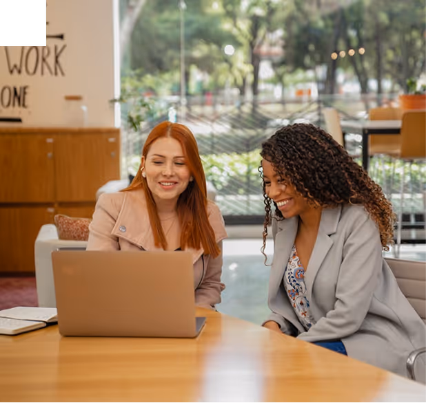 BBC - Deux femmes souriantes utilisant un ordinateur portable ensemble dans un bureau lumineux.