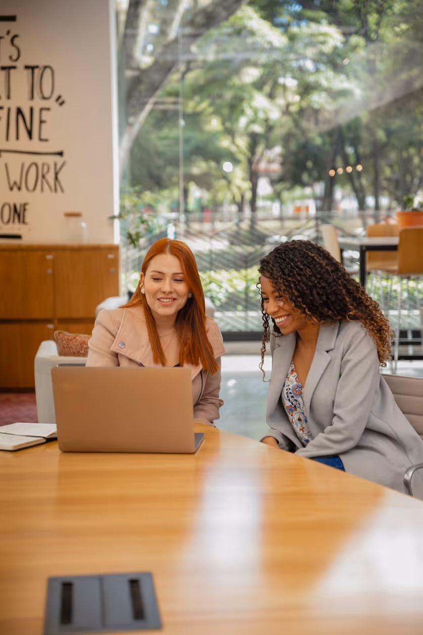 Deux femmes souriantes regardent un ordinateur portable posée sur une table en bois dans un bureau.