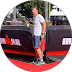 Homme en short blanc et t-shirt gris debout sur un tapis rouge devant un mur avec logos Ironman.